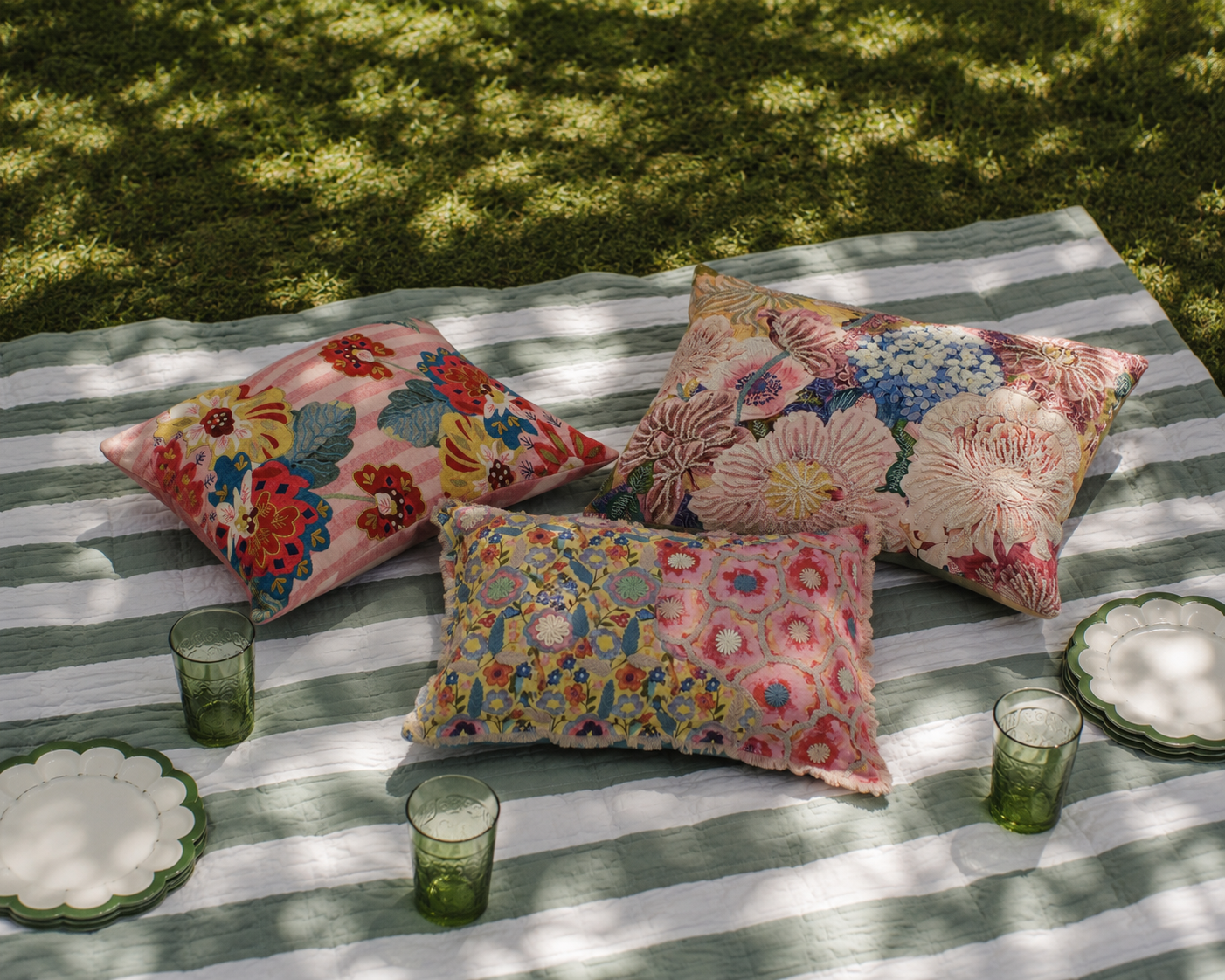 Colorful floral cushions on a striped blanket with glasses and plates outdoors.